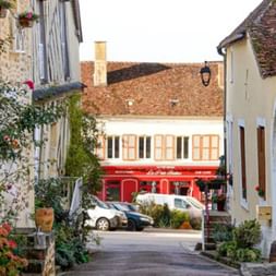 Enge Straße in Saint-Sauveur-en-Puisaye mit traditionellen Gebäuden, blühenden Pflanzen und einem roten Geschäft im Hintergrund.