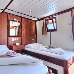 Zweibettkabine auf der MY Andela Lora mit zwei Einzelbetten, weißer Bettwäsche, Holzverkleidung und großem Fenster mit Küstenblick.