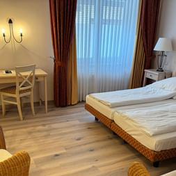 Helles Hotelzimmer im Hotel Central Zeven mit Doppelbett, Holzschreibtisch, Korbstühlen, Flachbildfernseher und roten Vorhängen.