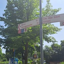 Zwei Radwegweiser an einem Mast zeigen Routen nach Bonn-Beuel/Vilich-winter und Unkel. Bäume und Wohnhäuser im Hintergrund.