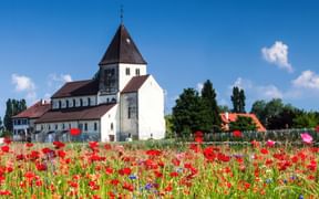 Historische St. Georg Kirche auf der Reichenau mit weißen Mauern und dunklem Kegeldach, umgeben von bunter Wildblumenwiese mit roten Mohn.