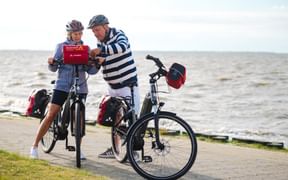 Two cyclists with helmets examining a red folder by the waterfront. Their touring bikes with red panniers are parked on a paved path beside the sea.