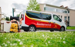 White and red Rückenwind van parked on grass with suitcases and yellow backpack beside it. Residential buildings visible in background.