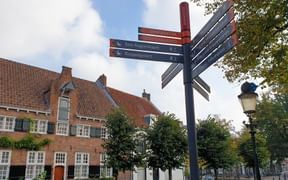 Mehrfach-Wegweiser mit farbigen Pfeilen zu verschiedenen Zielen auf einem Stadtplatz mit historischen Backsteingebäuden.