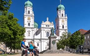 Passauer Dom mit zwei barocken Türmen mit grünen Zwiebelkuppeln. Zwei Radfahrer mit Rädern stehen auf dem Platz vor der weißen Fassade.