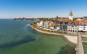 Luftaufnahme der Friedrichshafener Uferpromenade am Bodensee mit Gebäuden, Steg und Kirchturm unter blauem Himmel.