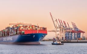 Großes COSCO-Containerschiff mit bunten Containern am Hamburger Hafenterminal mit Kränen und Schlepper bei Sonnenuntergang.