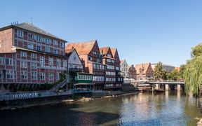 Traditionelle Fachwerkhäuser und Backsteingebäude säumen die Uferpromenade in Lüneburg, mit einem Fluss, der die bunten Fassaden unter blauem Himmel spiegelt.