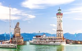 Lindauer Hafen am Bodensee mit weißem Leuchtturm, Löwenstatue, Passagierboot und Segelboot. Alpenkulisse im Hintergrund sichtbar.
