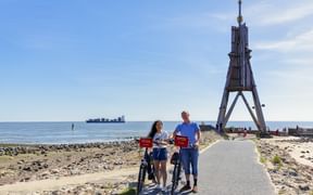 Zwei Radfahrer mit roten Packtaschen stehen mit ihren Rädern an der hölzernen Kugelbake in Cuxhaven, mit Nordsee und Schiff im Hintergrund.
