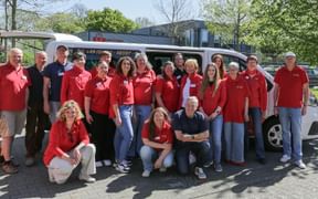 Large team group photo with people in red shirts standing and kneeling in front of a white van, outdoors on a sunny day.