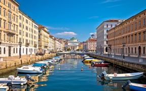 Canal Grande in Triest mit bunten Booten an Steinkai vertäut. Historische neoklassizistische Gebäude säumen beide Seiten unter blauem Himmel.
