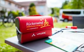 Red VAUDE handlebar bag with Rückenwind cycling tour branding placed on a table with maps and documents, bicycles visible in background.