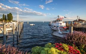 Hafen in Unteruhldingen am Bodensee mit Booten, Holzstegen und bunten Blumen im Vordergrund unter blauem Himmel mit Wolken.