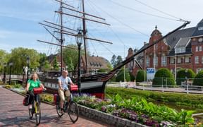 Zwei Radfahrer mit roten Packtaschen neben einem historischen Segelschiff mit drei Masten in Papenburg. Traditionelle Backsteingebäude und bunte Blumen sichtbar.