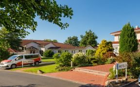 White hotel buildings with red tile roofs in Auefeld, surrounded by green lawn and colorful garden. Hotel shuttle van parked in front.