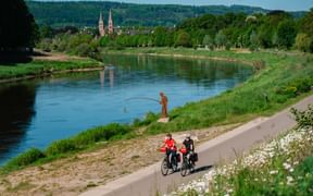 Zwei Radfahrer auf befestigtem Weg entlang der Weser mit Angler am Flussufer. Historisches Höxter mit Doppeltürmen im Hintergrund.
