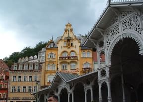 Weiße verzierte Kolonnade mit dekorativen Bögen im Vordergrund, Hotel Romance als gelbes Barockgebäude mit Turm in Karlsbad sichtbar.