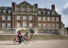 Two cyclists with touring bikes ride past a grand baroque castle with red brick facade and ornate central pavilion in Münsterland, Germany.