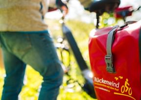 Red cycling panniers with Rückenwind branding attached to a bicycle, with cyclists' legs visible in the background on a sunny day.
