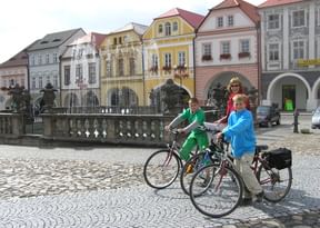 Drei Radfahrer mit Rädern posieren an einem verzierten Brunnen auf dem historischen Marktplatz von Kadan, umgeben von bunten Barockhäusern.