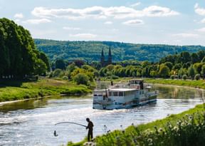 White river cruise ship sailing on calm river with green hills and church spires in background. Angler fishing from grassy riverbank.