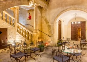 Elegant stone courtyard with arched architecture, wrought iron dining tables and chairs, warm lighting, and a grand staircase with balustrade.