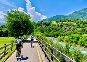 Two cyclists riding on a wooden bridge along the Etsch Valley bike path, surrounded by lush green mountains and turquoise river below.