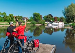 Zwei Radfahrer mit roten Taschen stehen auf einem Holzsteg und winken einem weißen Flusskreuzfahrtschiff zu. Der ruhige Fluss spiegelt Bäume und Häuser.