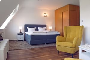 Helles Hotelzimmer mit dunkelblauem Boxspringbett, gelbem Ohrensessel, Holzboden und Dachschräge mit Dachfenster.