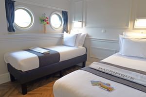 Helle Kabine auf der MS Magnifique Vier mit drei Einzelbetten, weißer Bettwäsche, Bullaugen mit blauen Vorhängen und Holzboden.