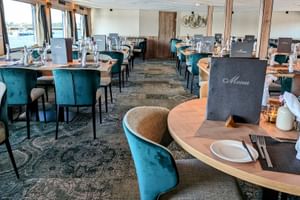 MS Princess Schiffsrestaurant mit runden Tischen, türkisfarbenen Samtstühlen und Gedecken. Menükarten auf Tischen, Fenster zeigen Wasserblick.