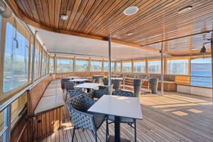 Geräumiges Deck der MY Magellan mit Holzdecke, weißen Bänken, Esstischen mit blauen Stühlen und großen Fenstern mit Blick auf die kroatische Küste.