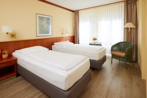 Helles Hotelzimmer mit zwei Einzelbetten, Holzkopfteil, grünem Sessel, Stehlampe und großem Fenster mit Gardinen im Hotel Geheimer Rat.