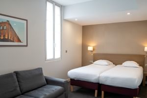 Modernes Hotelzimmer in Toulouse mit zwei Einzelbetten, grauem Sofa, Wandlampen und gerahmtem Kunstwerk mit historischem Gebäude.