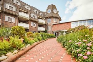 Backsteinhotel mit Turm und Balkonen in Varel. Gepflasterter Weg führt durch Garten mit bunten Blumen und grünen Pflanzen.
