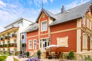 Orangefarbenes historisches Gebäude mit cremefarbenen Verzierungen und grauem Dach, Parkhotel Jever. Modernes weißes Hotelgebäude dahinter.