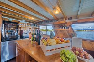 Innenraum des Linda Schiffssalons mit Holztheke, frischen Früchten in Holzkisten, Weinflaschen und Küchengeräten. Große Fenster zeigen Wasserblick.