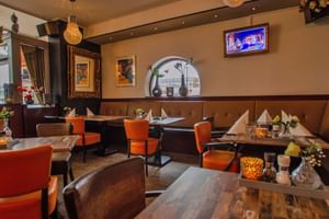 Gemütliche Hotelbar mit orangen Stühlen, Holztischen, braunen Sitzbänken und dekorativer Wandkunst. Ein TV-Bildschirm und Rundbogen sind sichtbar.