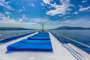 Sonnendeck des Schiffes Linda mit Reihen blauer gepolsterter Liegen auf weißem Deck, Metallgeländer und malerischem Seeblick mit Bergen.