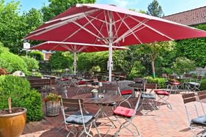 Außenterrasse im Landhotel Schnuck mit Ziegelpflaster, Holztischen, Metallstühlen mit Kissen und großem roten Sonnenschirm. Grüner Garten rundherum.