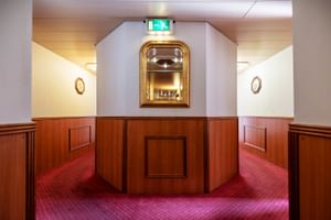 Schiffskorridor auf der MS Olympia mit rotem Teppich, holzgetäfelten Wänden und dekorativem Messingsspiegel in der Mitte. Notausgangsschild sichtbar.