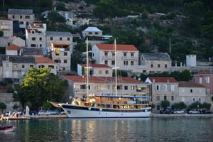 Weißes Passagierschiff San Snova mit zwei Masten vor Anker in ruhigen Hafengewässern. Steingebäude mit roten Ziegeldächern säumen den Hang.