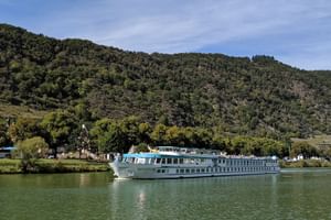 MS Switzerland Flusskreuzfahrtschiff auf ruhigem Wasser mit bewaldeten Bergen und kleinem Dorf im Hintergrund unter blauem Himmel.