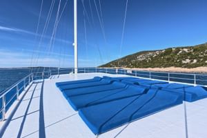 Sonnendeck des Schiffes San Snova mit blauen Liegen, weißem Mast und Reling. Kroatische Küste mit Hügeln im Hintergrund.