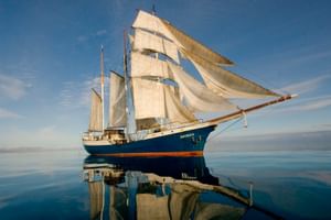 Das Segelschiff Antigua mit weißen Segeln gleitet über spiegelglattes blaues Wasser unter klarem Himmel und erzeugt eine perfekte Spiegelung.