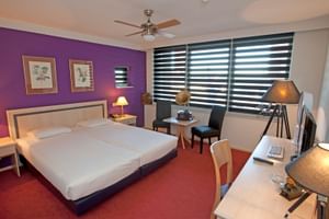Helles Hotelzimmer mit lila Akzentwand, Doppelbett, rotem Teppich, Schreibtisch, Sitzbereich und großen Fenstern mit Streifenjalousien.