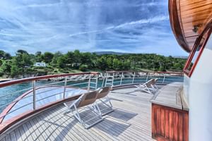 Sonnendeck der Andela Lora mit Holzboden, Liegestühlen und Metallgeländer mit Blick auf türkisfarbenes Wasser und bewaldete Küste.