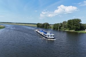 Weißes Flusskreuzfahrtschiff MS Princess mit blauen Akzenten fährt auf breitem Fluss unter blauem Himmel mit Wolken. Grüne Ufer und Bäume sichtbar.