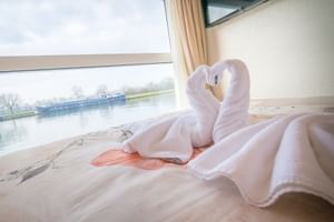 Kabineninterieur auf MS Fluvius mit schwanenförmigem Handtuch auf dem Bett, großes Fenster mit Flussblick und vorbeifahrendem Frachtschiff.
